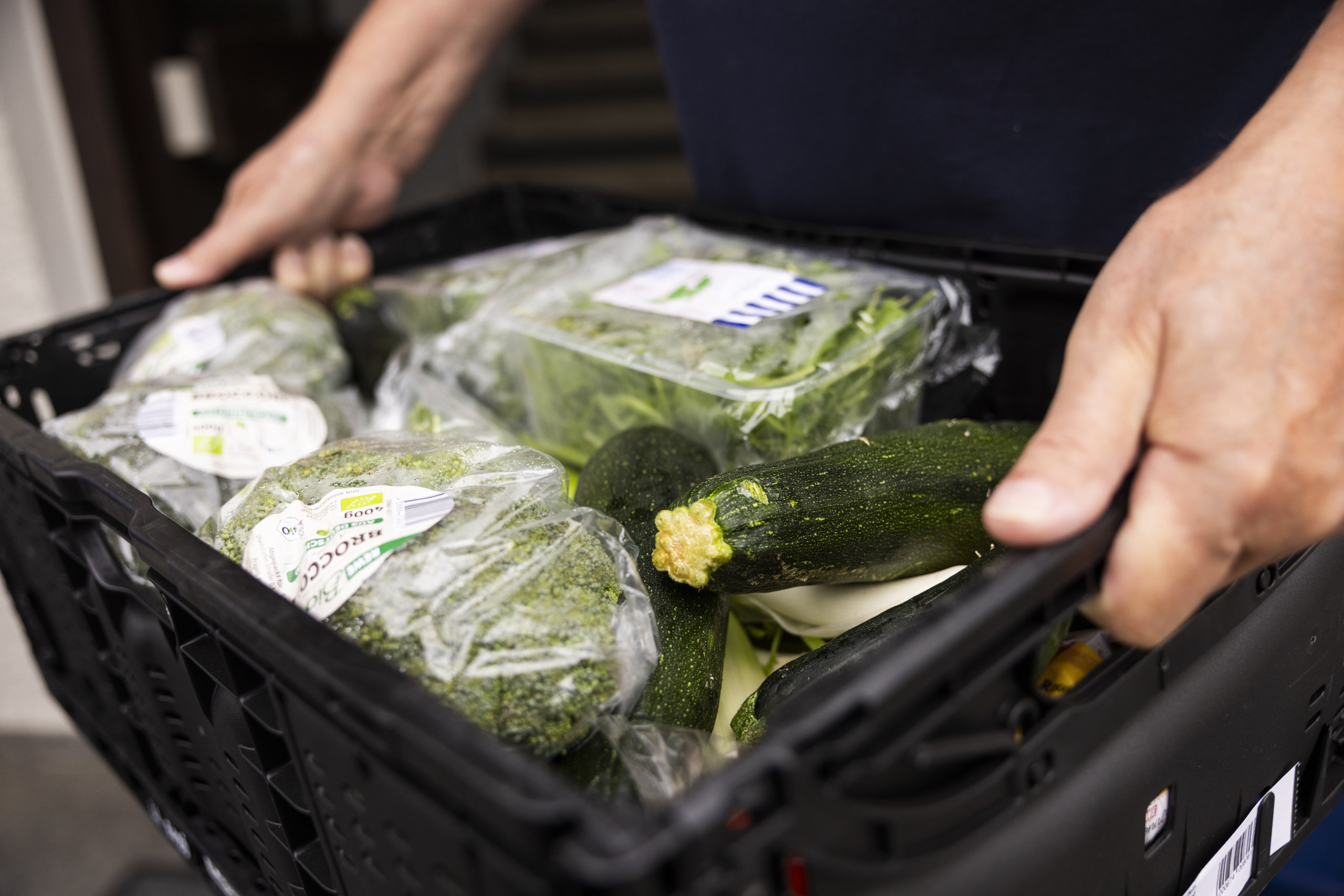 Nahaufnahme von frischem Gemüse, darunter Zucchini und Brokkoli, in einer schwarzen Kiste, die von einem Helfer der Tettnanger Tafel getragen wird