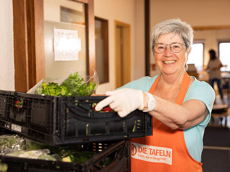 Eine lächelnde ehrenamtliche Helferin der Tettnanger Tafel trägt eine Kiste mit frischen Lebensmitteln, während sie eine Schürze mit dem Logo der Tafel trägt.