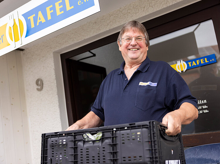 Ein freundlicher, älterer Mann, ein ehrenamtlicher Helfer, steht lächelnd mit einer Kiste vor der Tettnanger Tafel.