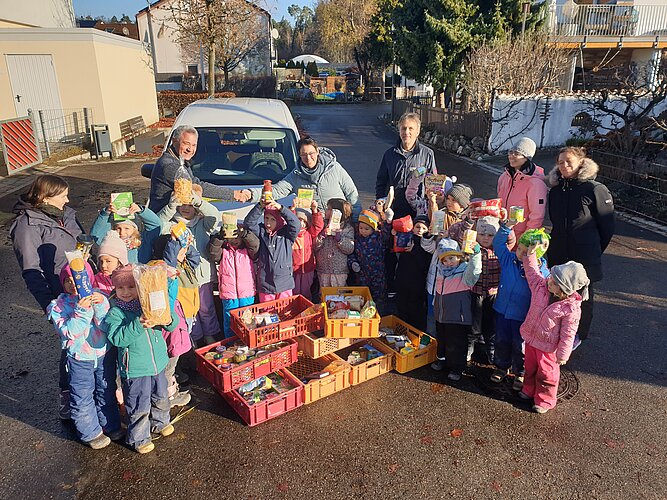 Spende des Kindergartens Bierkeller-Waldeck an die Tettnanger Tafel. Die Kinder vom Kindergarten stehen vor Kisten, gefüllt mit Spenden und halten Spenden hoch. Sie stehen vor Mitarbeitenden der Tettnanger Tafel und dem Auto.