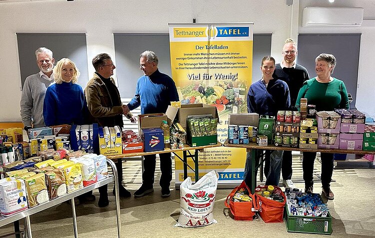 Klaus Nuber bedankt sich bei human aktiv e.V. im Tettnanger Tafelladen. Mehrere Personen stehen vor Bänken mit Lebensmittelspenden in der Tettnanger Tafel und lächeln in die Kamera.