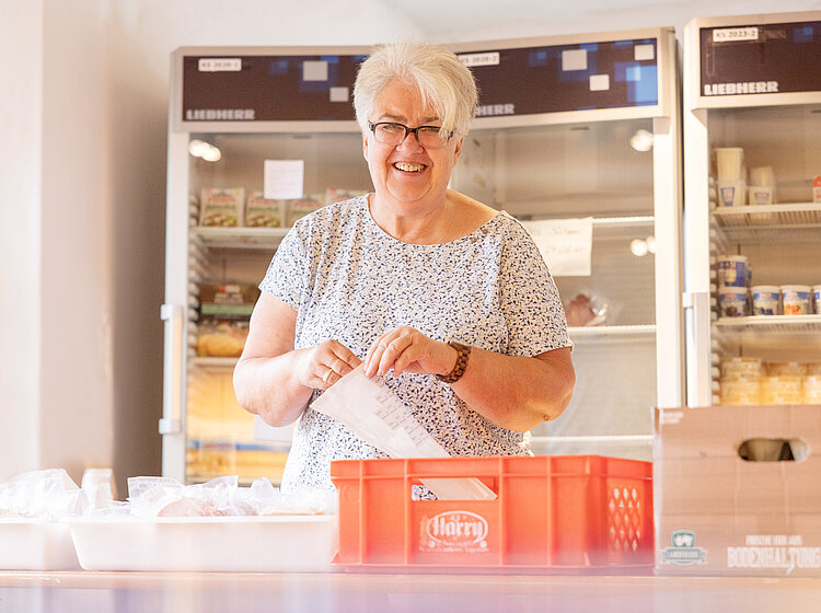 Eine lächelnde ehrenamtliche Helferin der Tettnanger Tafel verpackt Lebensmittel in einem Raum mit Kühlschränken im Hintergrund.