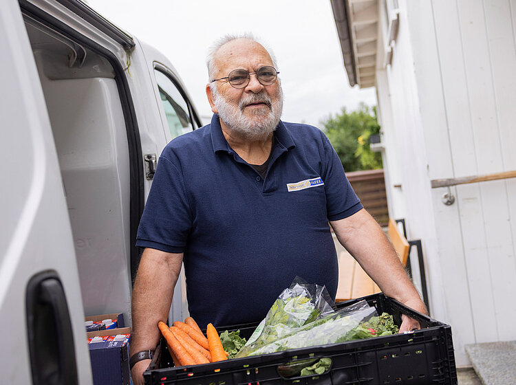 Ein ehrenamtlicher Helfer der Tettnanger Tafel steht mit einer Kiste voller frischem Gemüse neben einem Lieferwagen.